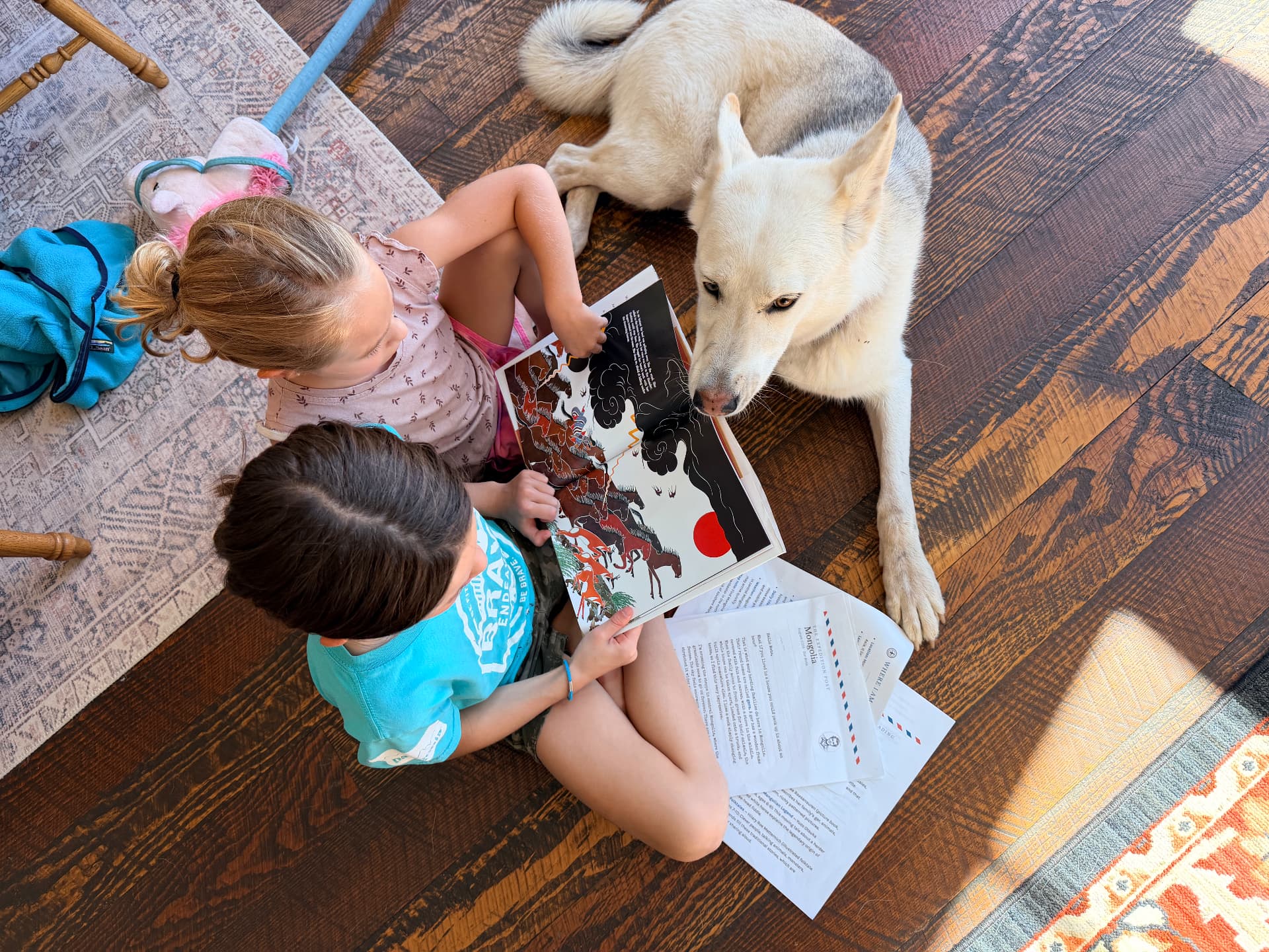 Two kids reading a picture book together on the living room floor with their dog, Rex's letter from Mongolia spread out beside them